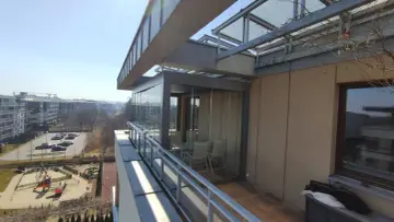 Nowoczesny ogród zimowy z przeszklonymi ścianami, stołem i krzesłami, oferujący widok na osiedle i plac zabaw.