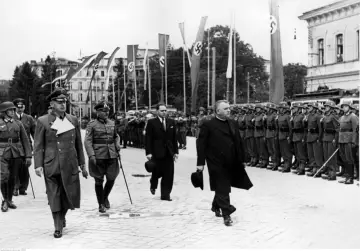 Urzędnicy w mundurach i ksiądz idą w defiladzie, w tle flagi z swastykami. Orawa.