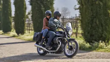 Coppia su una Moto Guzzi V7, con passeggero, percorre una strada alberata in una giornata di sole.