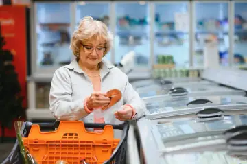 Starsza pani w sklepie analizuje cenę ryby, zastanawiając się nad skutkami wprowadzenia podatku od sprzedaży detalicznej dla konsumenta.