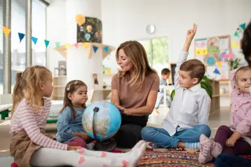 Nauczycielka z globusem w przedszkolu. Dzieci słuchają, jedno podnosi rękę. Ciekawe, ile się płaci za przedszkole?