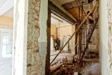 Trwa częściowe wyburzenie ściany nośnej. Widać robotników, rusztowania i gruz.