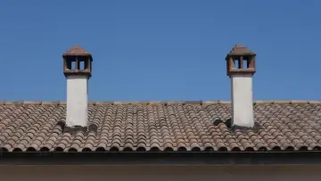 Dwa kominy na tle błękitnego nieba, osadzone na ceramicznym opierzeniu dachu.