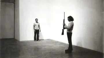 Chris Burden, dans une performance tendue, tient un fusil. Un homme se tient à distance, le regard fixé.
