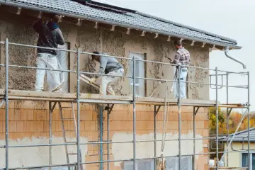 Pracownicy na rusztowaniu wykonują tynkowanie elewacji domu, nakładając zaprawę na ściany.