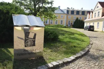Obok dworku w Soplicowie, na tle żółtego budynku, stoi metalowa księga z napisem "Podróże z Panem Tadeuszem".