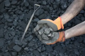 Ręce w rękawiczkach trzymające węgiel na tle stosu węgla i łopaty.