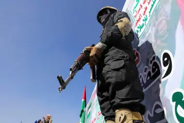 Mężczyzna w czarnym stroju i masce trzyma karabin. W tle transparent z arabskim napisem, symbolizujący ruch Huti.