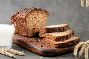 Kromka chleba proteinowego z ziarnami, obok szklanka mleka i kłosy zboża.