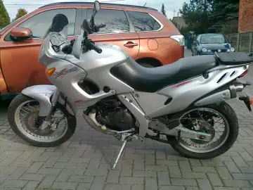 Srebrny motocykl Aprilia Pegaso 650 stoi na bruku obok pomarańczowego SUV-a.