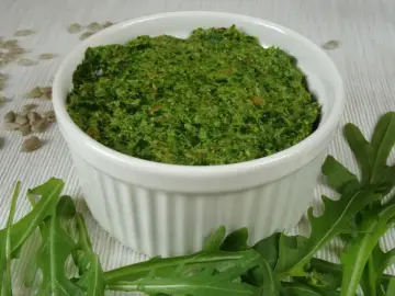Pesto z rukoli w białej miseczce, otoczone świeżymi liśćmi rukoli i nasionami słonecznika.