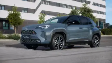 Szary Toyota Yaris Cross benzyna GR Sport z czarnymi dodatkami stoi na chodniku przed nowoczesnym budynkiem.