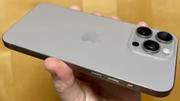 A hand holds a silver iPhone 15 Pro Max, showing its triple camera system and Apple logo.
