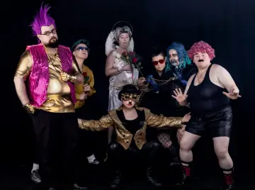 Grupa drag kingów w ekstrawaganckich strojach, z kolorowymi perukami i błyszczącymi dodatkami, pozuje do zdjęcia na czarnym tle.