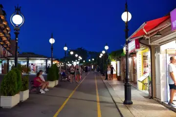 Wieczorna atmosfera na promenadzie w Złotych Piaskach. Ludzie spacerują, sklepy są otwarte, a latarnie rzucają ciepłe światło.