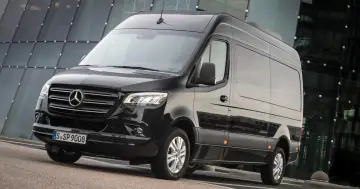 Czarny furgon Mercedes-Benz Sprinter stoi przed nowoczesnym budynkiem z przeszkloną fasadą.