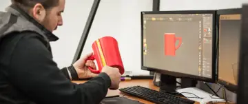 Mężczyzna mierzy czerwoną kubek 3d, porównując go z modelem na ekranie komputera.