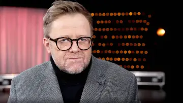 Olaf Lubaszenko w okularach, w szarym garniturze i czarnym golfie, na tle czerwonej kurtyny i świateł.