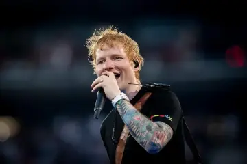 Ed Sheeran śpiewa z mikrofonem, jego ręka ozdobiona jest kolorowymi tatuażami.