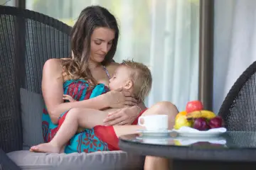 Matka karmi piersią dziecko na tarasie. Obok filiżanka kawy i owoce.