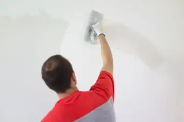 Mężczyzna w czerwonej bluzie nakłada gładź szpachlową na ścianę. Czas, ile schnie gładź szpachlowa, zależy od warunków.