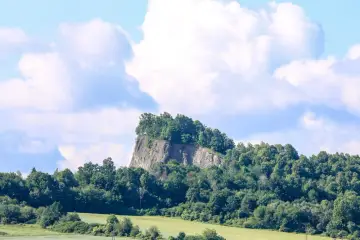 Skalisty szczyt porośnięty drzewami, przypominający wilczą górę w Złotoryi, góruje nad zielonymi polami i lasami.