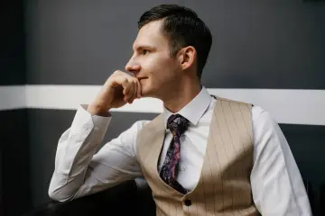 Elegancki mężczyzna w kamizelce, co zamiast marynarki, z dłonią na brodzie, wpatrzony w dal.