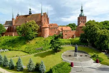 Zamek Frombork z katedrą i wieżą, otoczony zielenią. Na pierwszym planie pomnik na okrągłym placu.