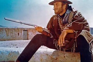 Clint Eastwood w wieku z karabinem, w ponczo i kapeluszu, z papierosem w ustach, siedzi na kamieniu.