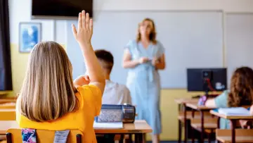 Uczennica z podniesioną ręką na lekcji. Czy po radzie pedagogicznej można zmienić oceny? Nauczycielka w tle.