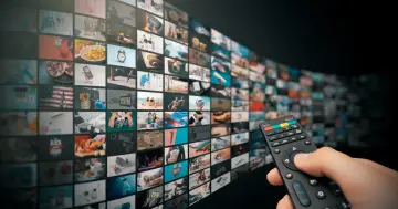 Dłoń trzyma pilota, wybierając treści z ekranu z wieloma oknami, jak w interfejsie **hbbtv**.
