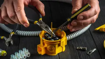 Ręce elektryka podczas osadzania puszek elektrycznych, używając szczypiec i śrubokręta do podłączenia przewodów.