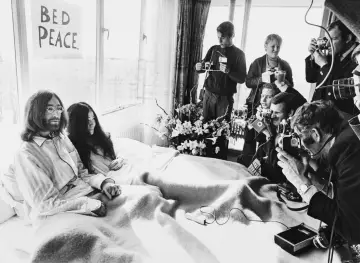 John Lennon i Yoko Ono w łóżku, otoczeni przez reporterów z aparatami. Na ścianie wisi plakat "BED PEACE".