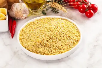 Miseczka pełna makaronu w kształcie ryżu, obok czosnek, papryczki chili, pomidorki i oliwa.