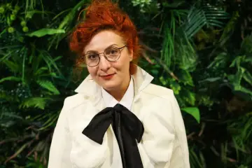 Kobieta z rudymi włosami i okularami, w białej marynarce z czarną kokardą, jak z obsady "Emmy".