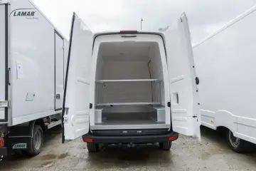 Biała izoterma z otwartymi drzwiami tylnymi, widoczne półki i wnętrze. Obok inne białe zabudowy.