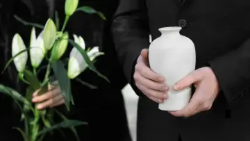 Urzędnik w czerni trzyma urnę, symbolizującą, co się dzieje z ciałem podczas kremacji. Obok białe lilie.