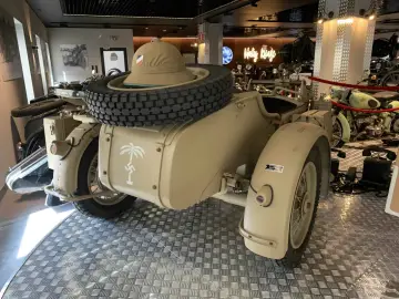 Piaskowy motocykl BMW R75 Sahara z przyczepką, kołem zapasowym i kapeluszem w tle.