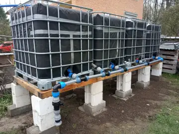 System zbiorników IBC, każdy z czarną osłoną, połączony rurami. Wygląda jak prowizoryczna instalacja do gromadzenia deszczówki, może nawet do nawadniania ogrodu, jak w przypadku systemu mauser.