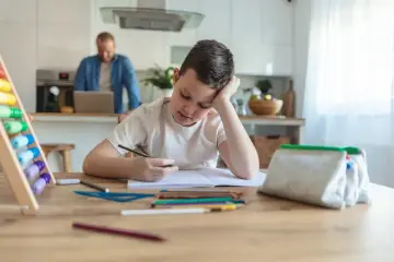 Chłopiec znużony odrabianiem lekcji, marzy o dniu bez prac domowych. W tle ojciec pracuje przy laptopie.