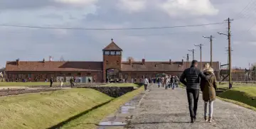 Para zwiedzająca Auschwitz indywidualnie idzie w stronę bramy obozu. W tle ceglane budynki i wieża strażnicza.