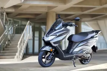 Szary skuter Suzuki Burgman 125 na tle nowoczesnego budynku z betonowymi schodami.
