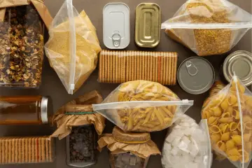 Zapasy żywności: słoiki z miodem, kawą, granolą, cukrem, paczki z makaronem, kaszą, muszelkami, puszki i herbatniki.