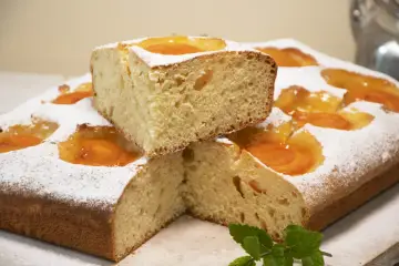 Deliciosa coca de albaricoque, cubierta de azúcar glas y decorada con trozos de fruta. Un trozo apilado revela su esponjosa miga.