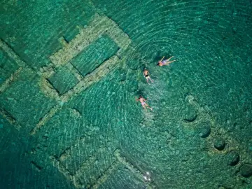 Swimmers explore the sunken city of Epidaurus, its ancient stone structures visible beneath the clear turquoise water.