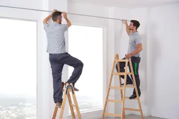 Zwei Männer auf Leitern bringen eine Gardinenschiene an.