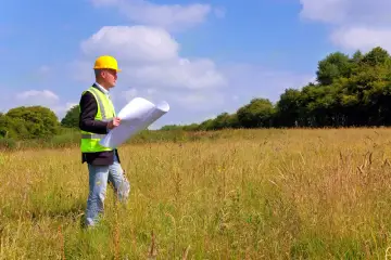 Mężczyzna w kasku i kamizelce odblaskowej analizuje plany na tle łąki. To pokazuje, co to znaczy nieruchomość gruntowa – potencjał do zagospodarowania.