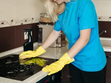 Kobieta w żółtych rękawiczkach czyści płytę ceramiczną, aby usunąć zapach spalonego mleka z płyty ceramicznej bez rysowania szkła.