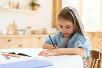 Dziewczynka z długimi włosami skupiona na pisaniu. Czy dysleksja to choroba? Pomocne mogą być kolorowe kredki i wsparcie.
