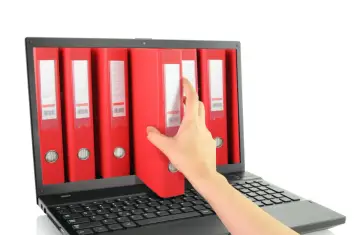 Dłoń sięga po czerwoną teczkę z cyfrowego archiwum na ekranie laptopa. To symbol cyfryzacji dokumentacji, np. w ramach zfśs.